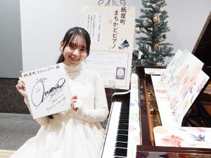 写真：サイン色紙を持ってピアノに座るまなまるさん